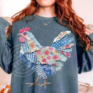 May include: A blue-grey sweatshirt features a patchwork rooster design. The rooster is made of various patterned fabrics, including floral, gingham, and polka dots, in shades of blue, pink, and white. The rooster's legs are a gold color.