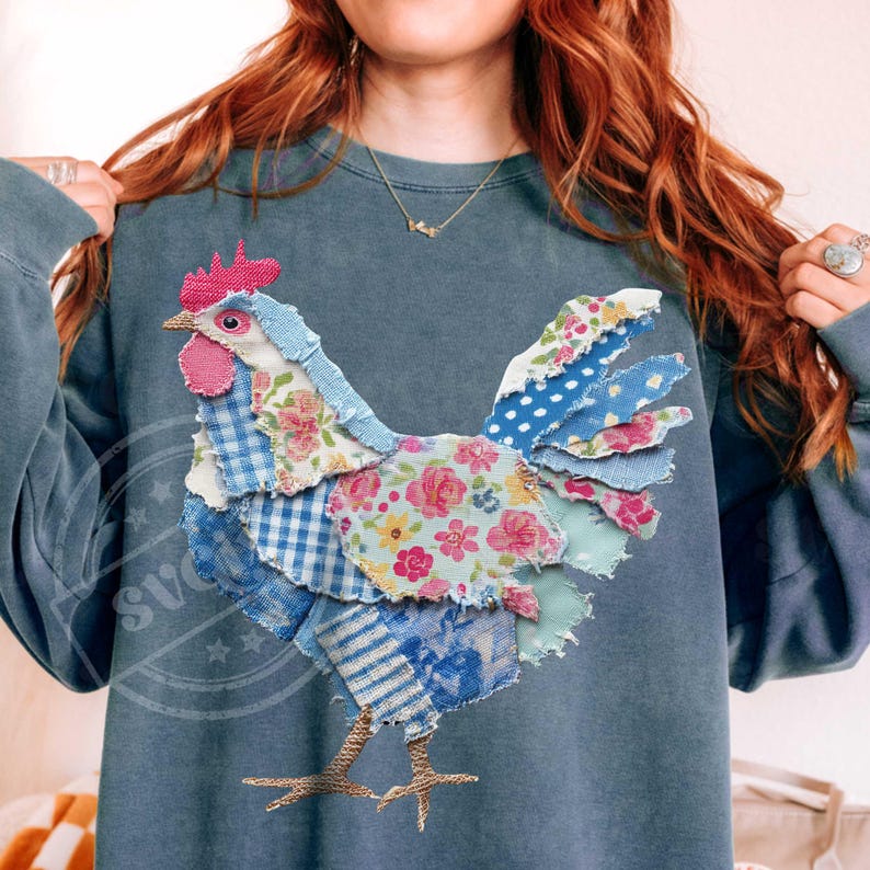 May include: A blue-grey sweatshirt features a patchwork rooster design. The rooster is made of various patterned fabrics, including floral, gingham, and polka dots, in shades of blue, pink, and white. The rooster's legs are a gold color.