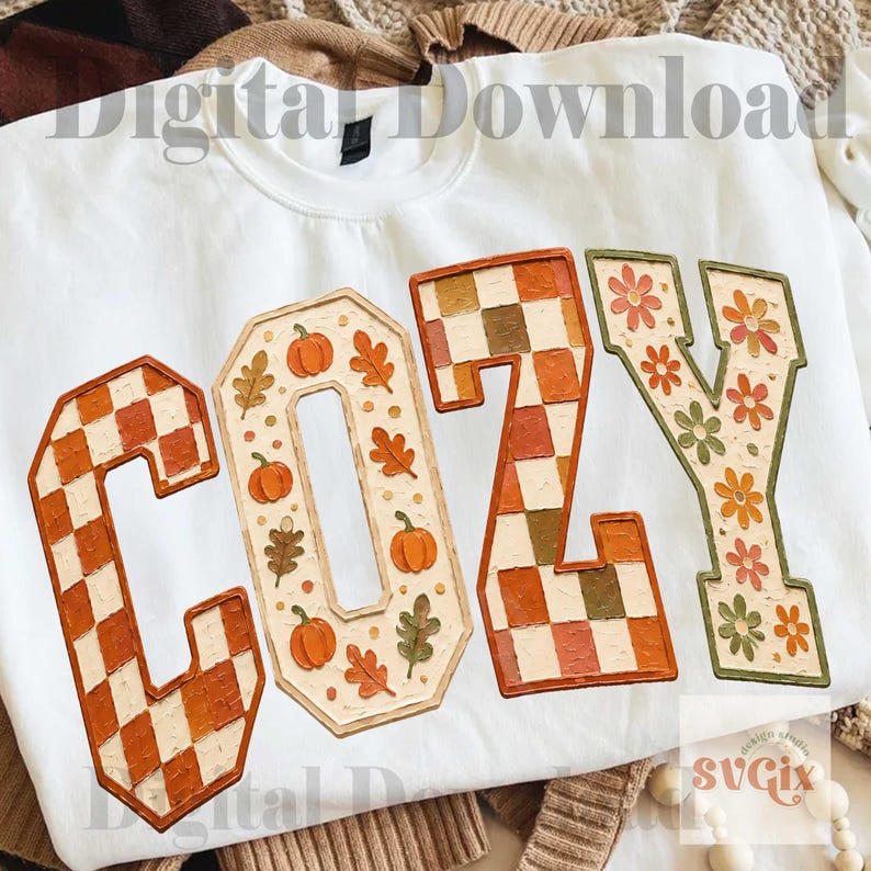 May include: A white sweatshirt featuring the word COZY in large block letters. The C and Z are filled with an orange and white checkerboard pattern. The O contains orange pumpkins and leaves, while the Y features orange and green flowers.