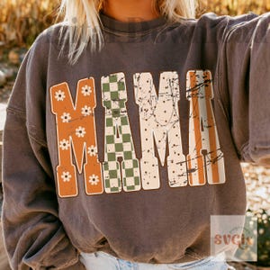 May include: A charcoal gray crewneck sweatshirt featuring the word MAMA in large block letters. The letters have a distressed texture and varied patterns: orange with white daisies, a green and white checkerboard, and cream with black dots.