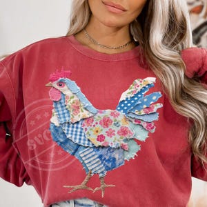 May include: A red sweatshirt featuring a patchwork rooster design. The rooster is made of various patterned fabrics, including floral, gingham, and polka dots. The rooster has a pink comb and wattle, and the sweatshirt has a crew neck.