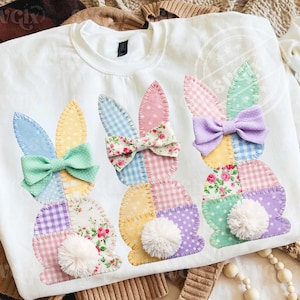 May include: White sweatshirt featuring three appliqued bunny designs. Each bunny has patchwork bodies, pom-pom tails, and patterned ears with a bow. The bows and ears are in pastel colors, including blue, pink, yellow, and purple, with floral and polka dot patterns.