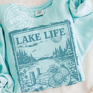 May include: A light blue sweatshirt with a screen-printed graphic that reads "LAKE LIFE." The graphic depicts a lake scene with trees, a dock, a hat, and sunglasses. The sweatshirt is folded and has a tag at the neckline.
