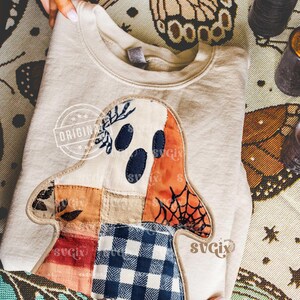 May include: Beige sweatshirt featuring a patchwork ghost design. The ghost has navy blue eyes, an orange spiderweb, and a patchwork of fabrics in various colors. The sweatshirt is folded and displayed on a patterned surface.
