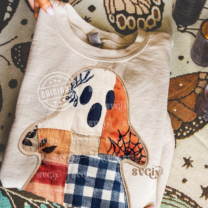 May include: Beige sweatshirt featuring a patchwork ghost design. The ghost has navy blue eyes, an orange spiderweb, and a patchwork of fabrics in various colors. The sweatshirt is folded and displayed on a patterned surface.