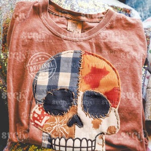 Puede incluir: Una camiseta de manga larga de color óxido con un diseño de calavera de patchwork. La calavera está hecha de varios patrones de tela, incluyendo cuadros azules, naranja y rojo. La camiseta tiene la palabra "ORIGINAL" impresa.