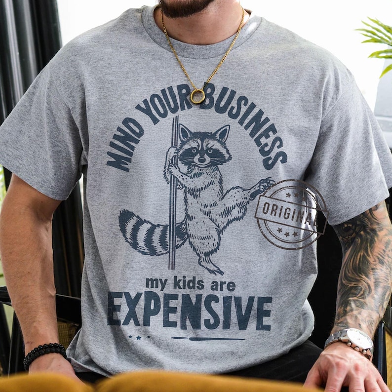 Pu&ograve; includere: T-shirt grigia con grafica di procione che tiene un palo, e la scritta "MIND YOUR BUSINESS" sopra il procione. Sotto il procione, la scritta recita "my kids are EXPENSIVE". Un timbro circolare "ORIGINAL" &egrave; a destra.