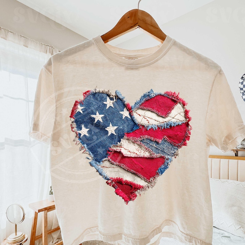 May include: A cream t-shirt featuring a heart-shaped design. The heart is crafted from denim and red fabric patches, with white stars on the blue denim, mirroring the American flag. The shirt is displayed on a wooden hanger.