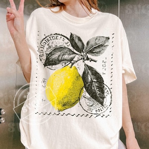 Könnte beinhalten: Cremefarbenes T-Shirt mit einem gelben Zitronen-Motiv und schwarzen Blättern, umgeben von einem Stempel-ähnlichen Design. Der Aufdruck enthält Text. Das T-Shirt hat kurze Ärmel und eine lockere Passform.