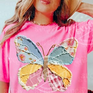 May include: A bright pink t-shirt featuring a colorful patchwork butterfly design. The butterfly has wings in shades of blue, yellow, pink, and red, with floral and plaid patterns. The shirt is a solid pink color.