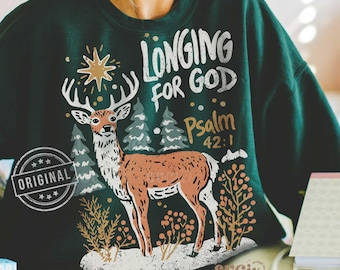 Puede incluir: Sudadera verde oscuro con un diseño de ciervo y el texto "LONGING FOR GOD Salmo 42:1." El ciervo es marrón y blanco, con cuernos, sobre un paisaje invernal con árboles y una estrella. La palabra "ORIGINAL" está estampada a la izquierda.