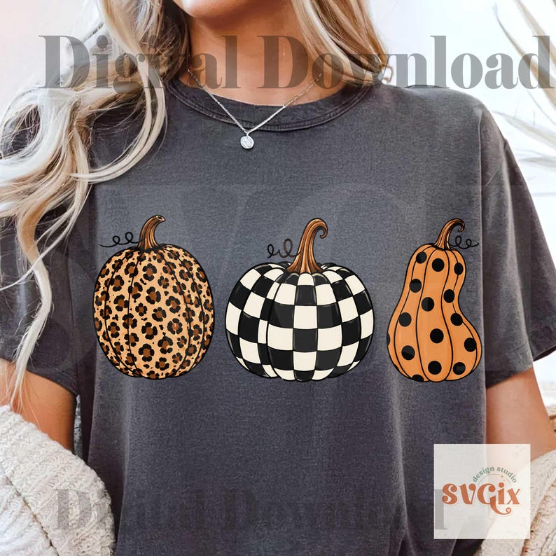 Puede incluir: Camiseta gris oscuro con tres ilustraciones de calabazas. Una calabaza tiene un estampado de leopardo, otra un patr&oacute;n de tablero de ajedrez en blanco y negro, y la tercera es naranja con lunares negros. La camiseta tiene el texto "Digital Download".