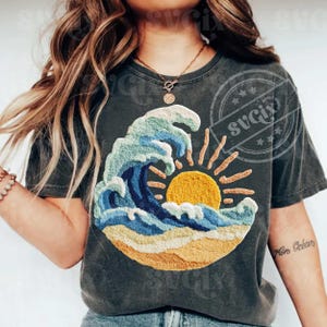 May include: Dark grey t-shirt with an embroidered design of a wave, sun, and beach scene. The wave is in blue and white, with a yellow sun and sandy beach. The shirt has a vintage, worn appearance.