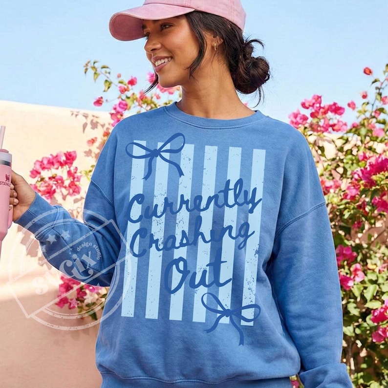 May include: A blue sweatshirt with vertical light blue stripes and the text "Currently Crashing Out" in a script font. The sweatshirt has a crew neck and long sleeves. A pink baseball cap is worn.