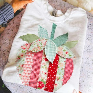 May include: White sweatshirt with an appliquéd strawberry design. The strawberry is made from various patterned fabrics in red, pink, and green. The leaves and stem are also fabric, adding texture. The design is a charming, handmade detail on the garment.