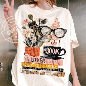 Puede incluir: Camiseta de color crema con un diseño de collage. El diseño incluye la palabra "BOOK" y una taza de café, gafas, flores y la palabra "LOVER" en una pila de libros. La camiseta tiene mangas cortas.