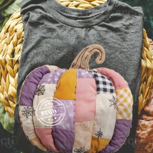 May include: A gray t-shirt with a decorative, patchwork pumpkin. The pumpkin is made of various fabrics in shades of purple, pink, yellow, and black and white. The pumpkin has a brown stem and spiderweb accents.