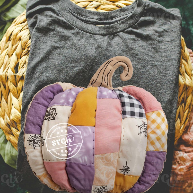 May include: A gray t-shirt with a decorative, patchwork pumpkin. The pumpkin is made of various fabrics in shades of purple, pink, yellow, and black and white. The pumpkin has a brown stem and spiderweb accents.