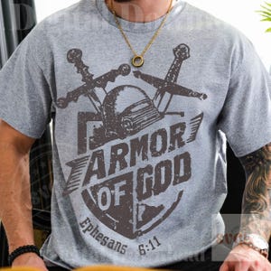 K&ouml;nnte beinhalten: Graues T-Shirt mit einem Schilddesign, das die Worte "ARMOR OF GOD" und den Text "Epheser 6:11" tr&auml;gt. Das Design zeigt einen Helm und gekreuzte Schwerter in Dunkelbraun.