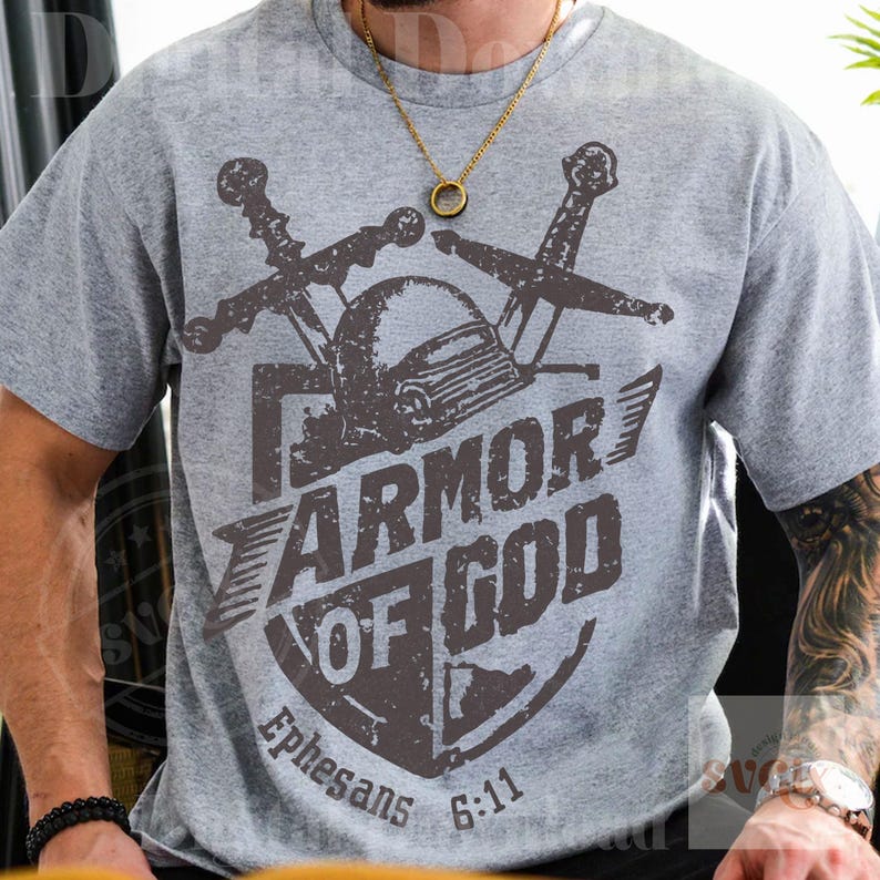 K&ouml;nnte beinhalten: Graues T-Shirt mit einem Schilddesign, das die Worte "ARMOR OF GOD" und den Text "Epheser 6:11" tr&auml;gt. Das Design zeigt einen Helm und gekreuzte Schwerter in Dunkelbraun.