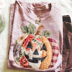 May include: Mauve t-shirt with a patchwork pumpkin design. The pumpkin has floral, polka dot, and plaid patterns, with a black face. A gold butterfly necklace rests on the shirt. The word "ORIGINAL" is stamped on the shirt.