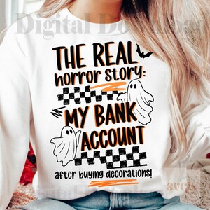 Pode incluir: Camisola branca com o texto "THE REAL horror story: MY BANK ACCOUNT after buying decorations!" em preto e laranja. Apresenta um morcego e duas ilustra&ccedil;&otilde;es de fantasmas, com um padr&atilde;o xadrez.