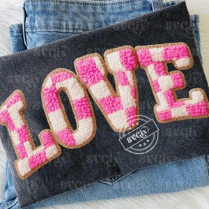 May include: A dark gray fabric with the word "LOVE" embroidered in pink and white checkered pattern. The letters have a fluffy texture and are outlined in a tan color. The fabric is resting on a pair of blue jeans.