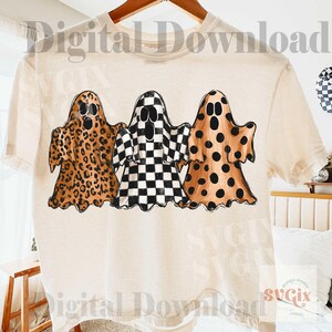 May include: A cream-colored t-shirt featuring a graphic of three ghosts standing side-by-side. The first ghost has a brown leopard print, the middle ghost has a black and white checkerboard pattern, and the third ghost has brown polka dots.
