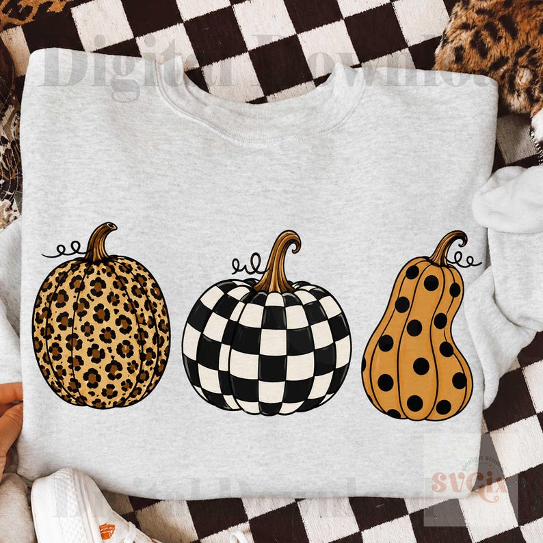 Puede incluir: Una sudadera gris claro con tres calabazas decorativas. Las calabazas tienen patrones &uacute;nicos: un estampado de leopardo, un tablero de ajedrez en blanco y negro y una calabaza naranja con lunares negros. Cada calabaza tiene un tallo marr&oacute;n y un zarcillo rizado.