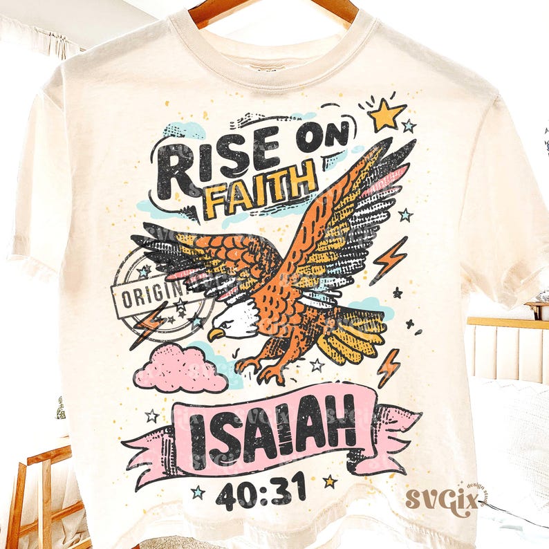Puede incluir: Camiseta color crema con un gr&aacute;fico de un &aacute;guila, las palabras "RISE ON FAITH" e "ISAIAH 40:31". El dise&ntilde;o incluye estrellas, rayos y una nube rosa. La camiseta est&aacute; colgada en una percha de madera.
