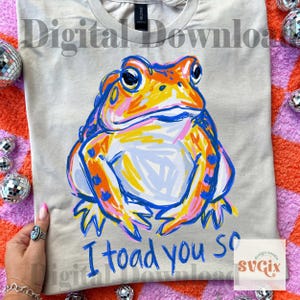 May include: A light gray t-shirt featuring a colorful illustration of a frog with orange, yellow, and blue accents. The text "I toad you so" is written in blue below the frog. The shirt is a digital download.
