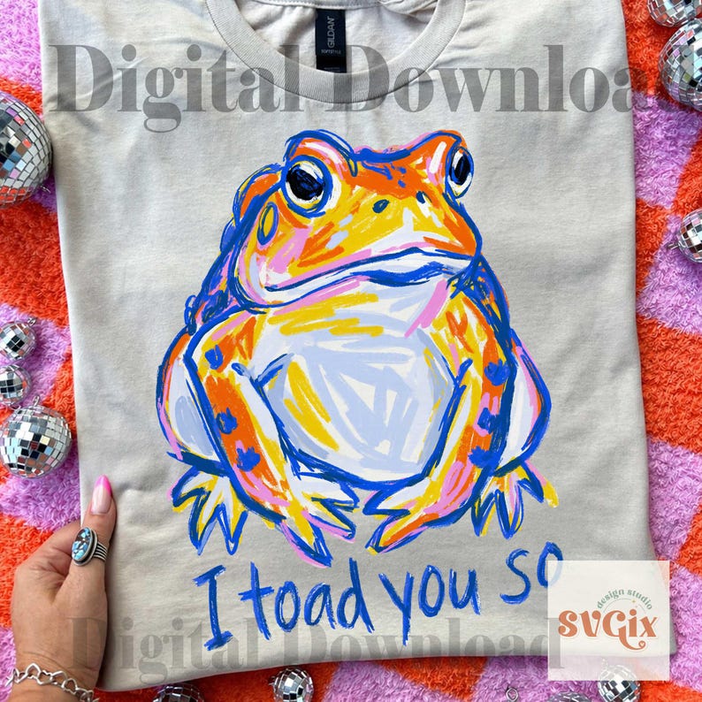 May include: A light gray t-shirt featuring a colorful illustration of a frog with orange, yellow, and blue accents. The text "I toad you so" is written in blue below the frog. The shirt is a digital download.