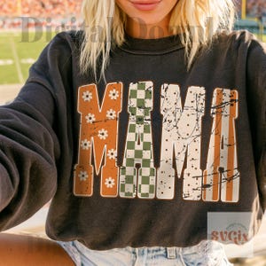 May include: A charcoal gray crewneck sweatshirt featuring the word MAMA in large block letters. Each letter has a distinct pattern: orange with white flowers, green and white checkerboard, white with black dots, and orange with white stripes.