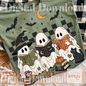 May include: An olive green t-shirt featuring a graphic of three white ghosts reading books. One ghost wears a black witch hat. The design includes a black cat, a pumpkin, a crescent moon, bats, stars, and a black checkered pattern background.