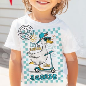 Pu&ograve; includere: Maglietta bianca con un motivo a scacchi blu e bianchi. Un'oca di cartone animato con occhiali da sole e un cappello blu sta guidando uno scooter. Il testo "On the Loose Like a Goose" &egrave; stampato sulla maglietta.