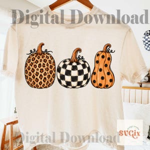 Puede incluir: Camiseta color crema con tres dise&ntilde;os de calabazas. Las calabazas presentan un estampado de leopardo, un patr&oacute;n de tablero de ajedrez en blanco y negro y una calabaza naranja con lunares negros. Las palabras "Digital Download" est&aacute;n impresas en la camiseta.
