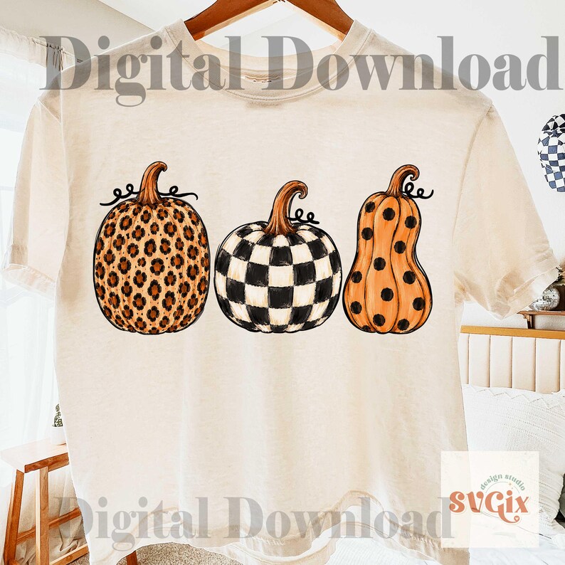 Puede incluir: Camiseta color crema con tres dise&ntilde;os de calabazas. Las calabazas presentan un estampado de leopardo, un patr&oacute;n de tablero de ajedrez en blanco y negro y una calabaza naranja con lunares negros. Las palabras "Digital Download" est&aacute;n impresas en la camiseta.