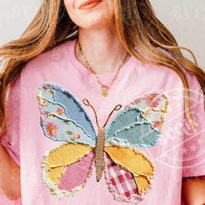 May include: Pink t-shirt featuring a large, patchwork butterfly design. The butterfly has wings in various colors and patterns, including floral, gingham, and solid fabrics. The shirt has a round neckline and short sleeves.