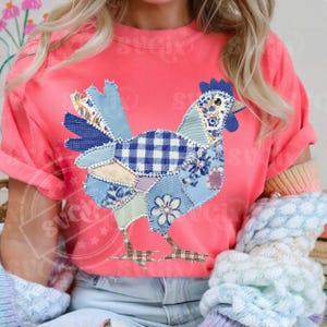 May include: A coral-colored t-shirt featuring a patchwork chicken design. The chicken is composed of various blue patterned fabrics, including gingham and floral prints. The shirt has rolled-up sleeves and is paired with light-wash jeans.