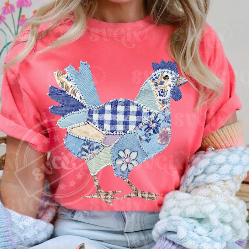 May include: A coral-colored t-shirt featuring a patchwork chicken design. The chicken is composed of various blue patterned fabrics, including gingham and floral prints. The shirt has rolled-up sleeves and is paired with light-wash jeans.