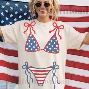 May include: Cream-colored t-shirt featuring a red and blue bikini design. The bikini top has a blue background with white stars, and the bottom has red and white stripes. The design evokes a patriotic theme.