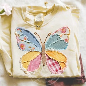 May include: Cream-colored long-sleeved shirt with a large, patchwork butterfly design. The butterfly features blue, pink, yellow, and red patterned fabrics. A gold butterfly necklace is draped on the shirt. The shirt is folded and appears to be made of soft material.