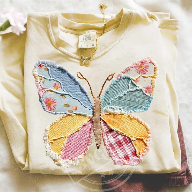 May include: Cream-colored long-sleeved shirt with a large, patchwork butterfly design. The butterfly features blue, pink, yellow, and red patterned fabrics. A gold butterfly necklace is draped on the shirt. The shirt is folded and appears to be made of soft material.