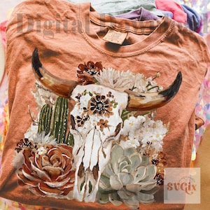 Puede incluir: Una camiseta de color naranja quemado con un estampado gr&aacute;fico de un cr&aacute;neo de vaca blanco adornado con flores marrones y blancas, cactus verdes y suculentas. El dise&ntilde;o est&aacute; centrado en el pecho y muestra una ilustraci&oacute;n de flora des&eacute;rtica.