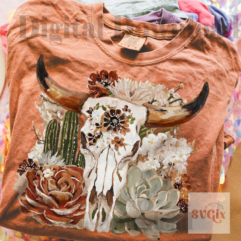 Puede incluir: Una camiseta de color naranja quemado con un estampado gr&aacute;fico de un cr&aacute;neo de vaca blanco adornado con flores marrones y blancas, cactus verdes y suculentas. El dise&ntilde;o est&aacute; centrado en el pecho y muestra una ilustraci&oacute;n de flora des&eacute;rtica.