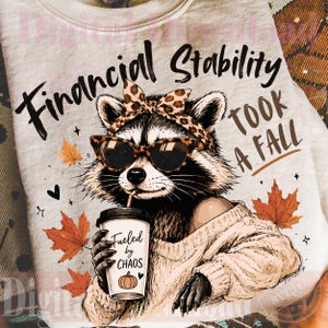 May include: A light-colored t-shirt featuring a graphic of a raccoon wearing a leopard print headband and sunglasses. The raccoon holds a coffee cup labeled "Fueled by CHAOS" with a pumpkin icon. Text reads "Financial Stability TOOK A FALL".
