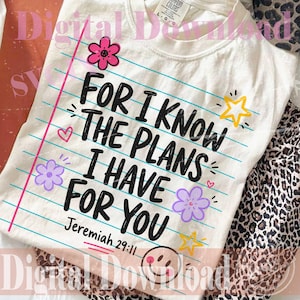 Può includere: Una t-shirt bianca con un design grafico che ricorda un foglio di quaderno con linee orizzontali blu e un margine verticale rosa. Il testo recita "FOR I KNOW THE PLANS I HAVE FOR YOU Jeremiah 29:11", accompagnato da fiori rosa e viola, stelle gialle e una faccina sorridente.