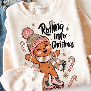 May include: Cream-coloured sweatshirt featuring a gingerbread person graphic wearing a pink and gold hat, scarf, and roller skates. The text "Rolling into Christmas" is above the gingerbread person, with candy canes and stars.