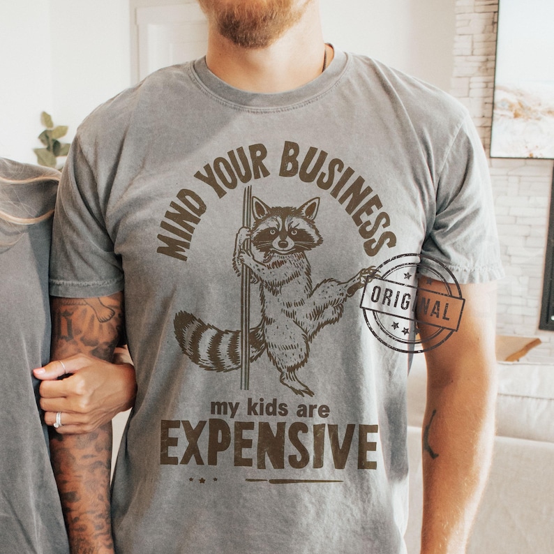 Pu&ograve; includere: T-shirt grigia con grafica di un procione. Il testo "MIND YOUR BUSINESS" &egrave; sopra il procione, con "my kids are EXPENSIVE" sotto. Un timbro circolare "ORIGINAL" &egrave; sul lato destro.