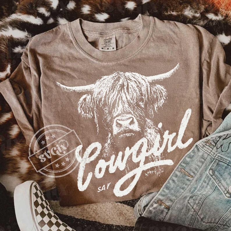 May include: A brown long-sleeve t-shirt featuring a white graphic of a Highland cow and the text "Cowgirl Say". The shirt is styled with a denim jacket and checkered shoes.